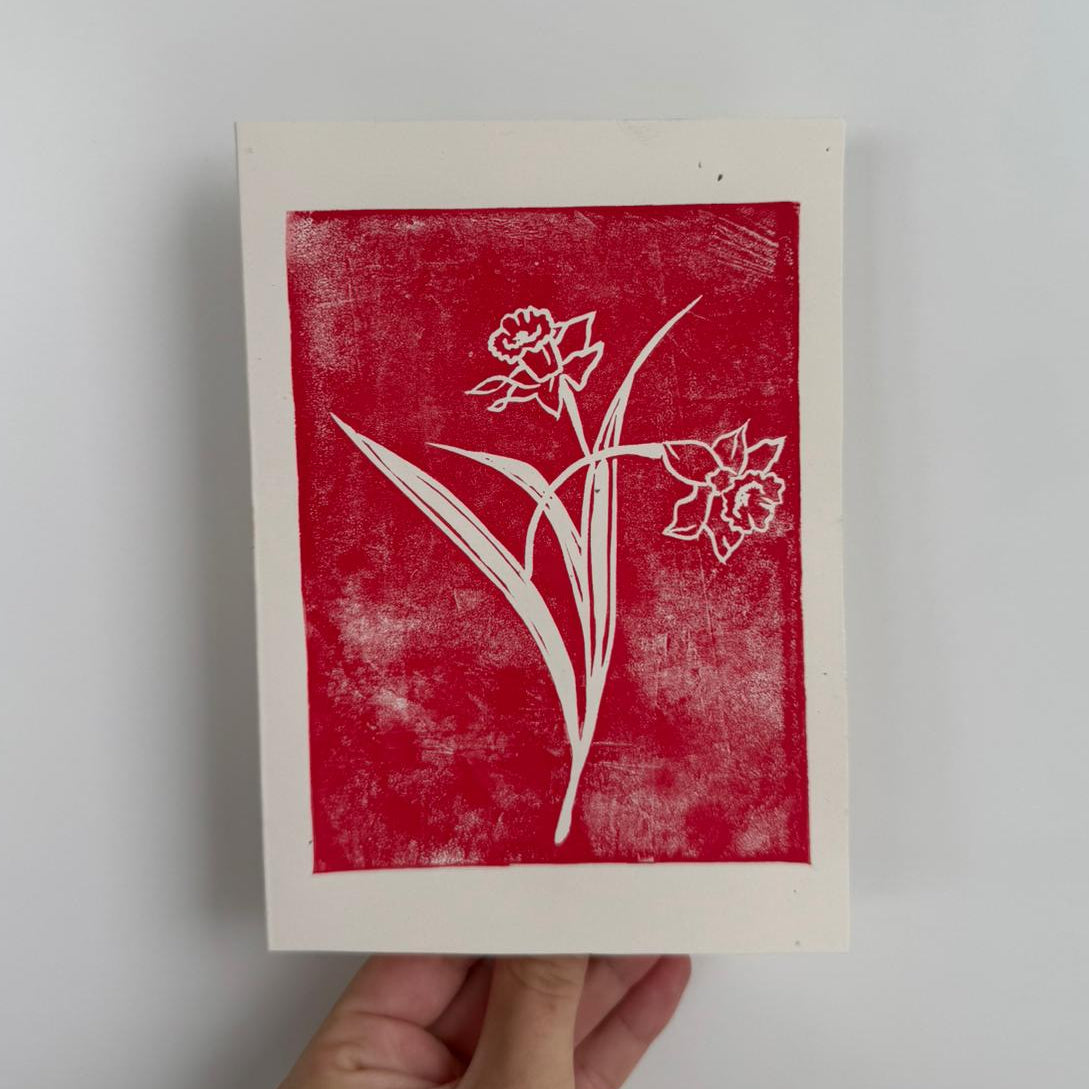 Handgemachte Linoldruckkarte mit rotem Motiv einer gelben Narzisse auf weissem Hintergrund. Zarte Blüte mit langen Blättern, minimalistisches Design im Linolschnittverfahren. Ideal als Frühlings- oder Osterkarte.