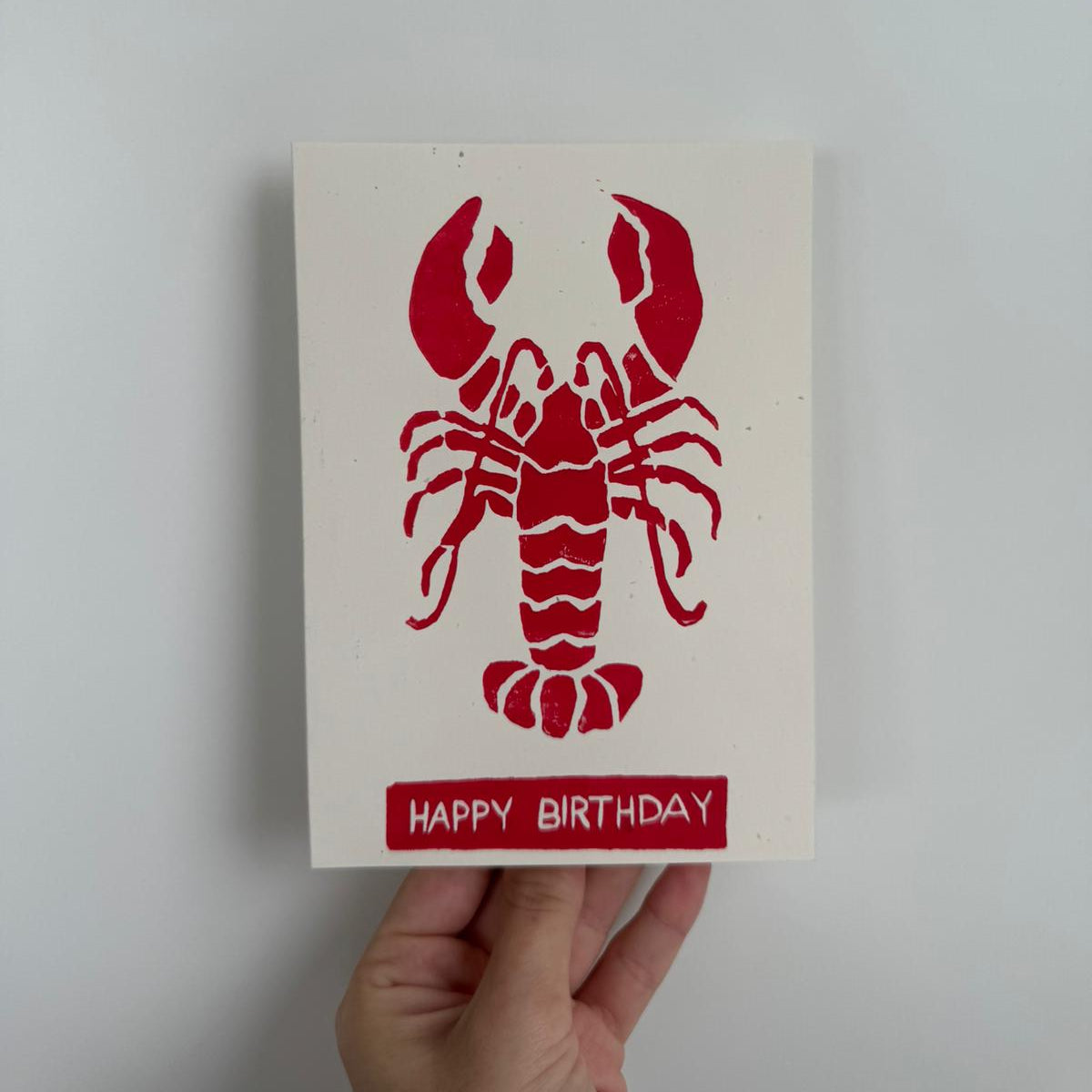 Handgemachte Linoldruckkarte mit rotem Hummer und der Aufschrift „Happy Birthday“. Minimalistisches Design auf naturweissem Hintergrund, handgedruckt im Linolschnittverfahren. Humorvolle und stilvolle Geburtstagskarte, jedes Stück ein Unikat.