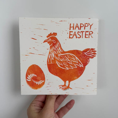 Handgemachte Linoldruckkarte mit orangefarbenem Huhn, Osterei und Schriftzug „Happy Easter“ auf weissem Hintergrund. Frühlingshaftes, verspieltes Motiv – ideal als Osterkarte oder Dekoration.